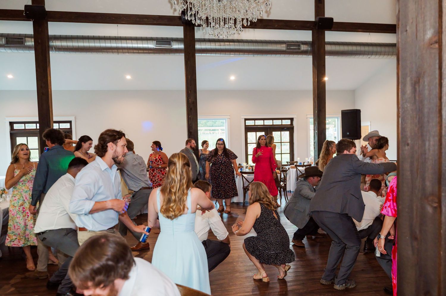 Guests in dressy attire dance and mingle on a wooden floor in a rustic event venue with exposed beams and a chandelier.