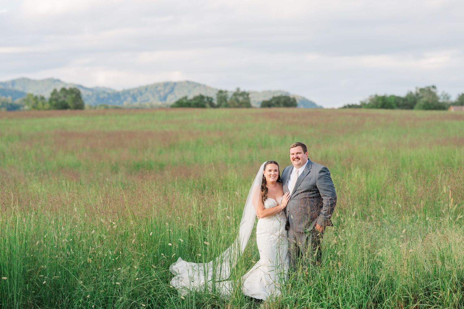 Bride and groom pose together in a tall grass field with rolling mountains in the background.