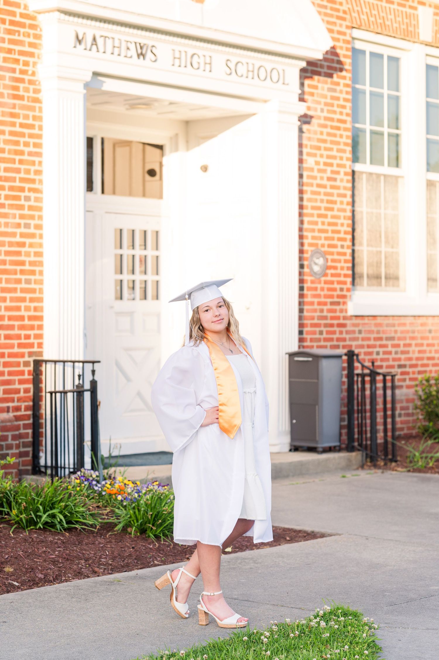 A graduate in white cap and gown poses playfully outside Mathews High School.