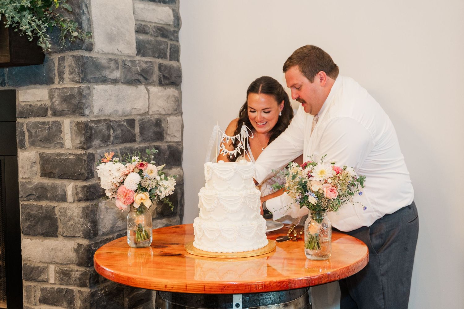 Bride and groom cut their white wedding cake together near a stone fireplace with floral arrangements nearby.