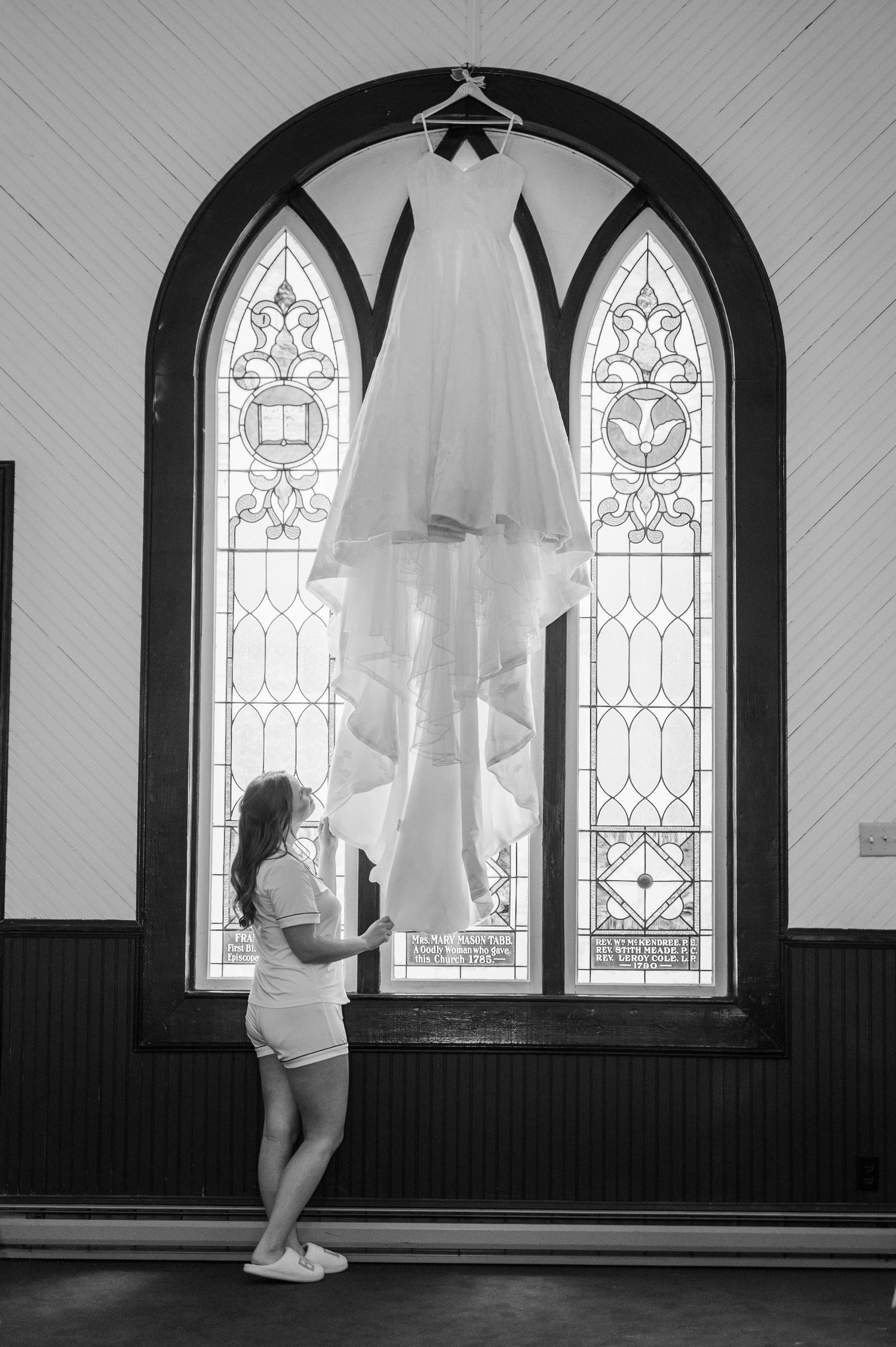 Black and white photo of a wedding dress hanging in front of gothic church windows.