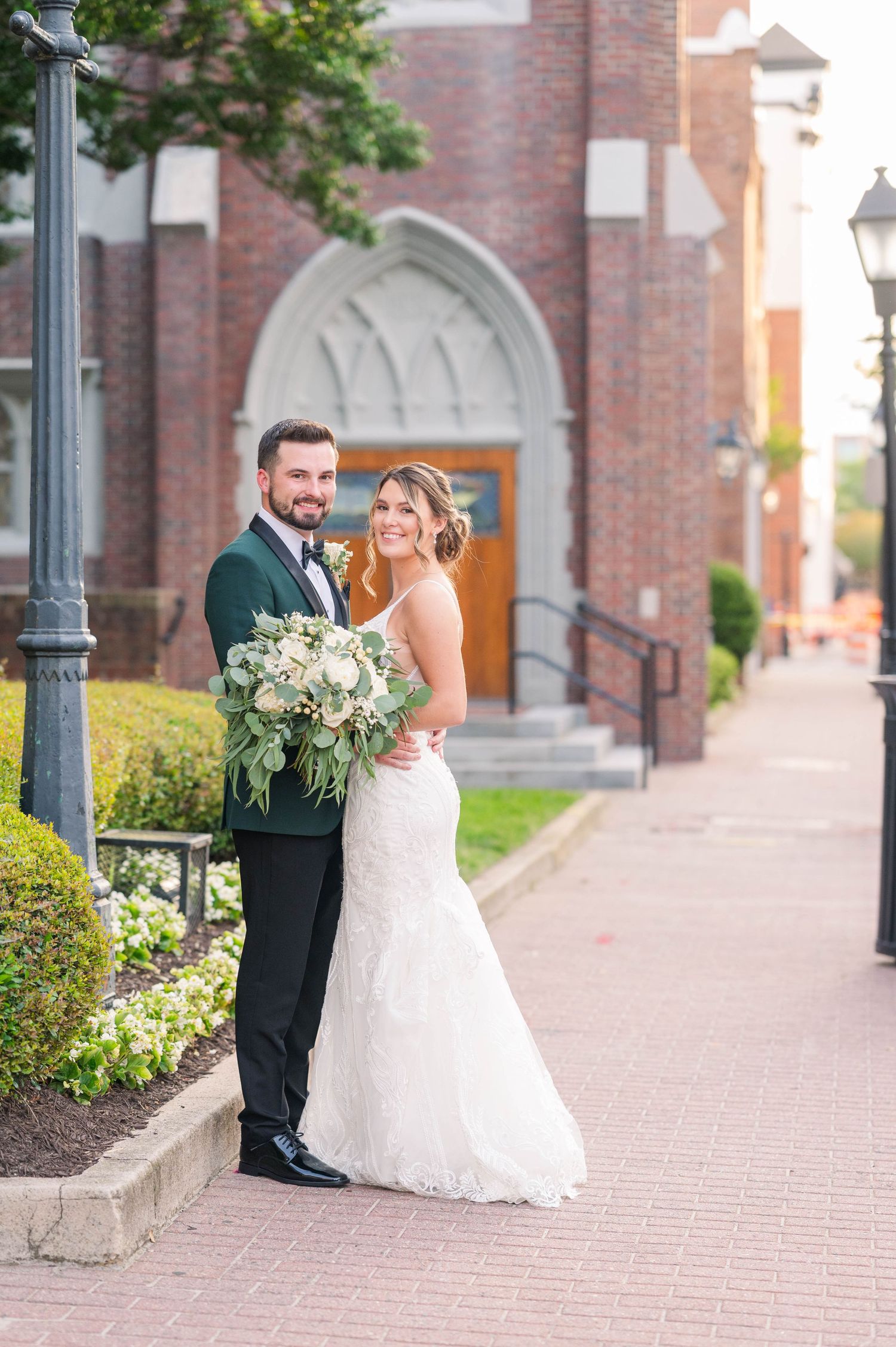 Wedding portraits along brick building exterior with vintage lamp posts and landscaping.