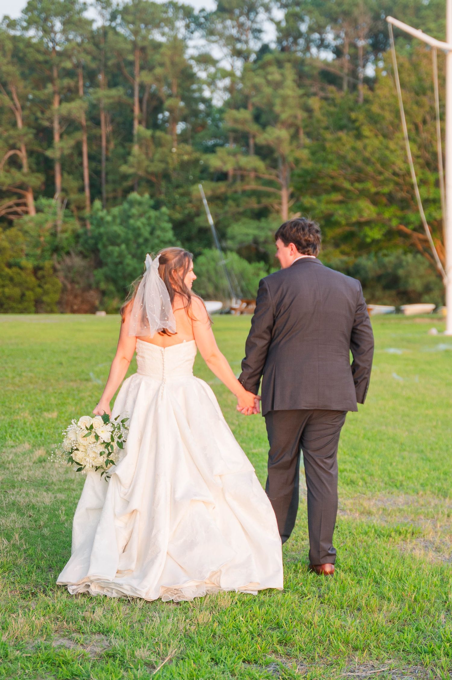 Wedding couple walks hand in hand across a grassy field with pine trees in the background.