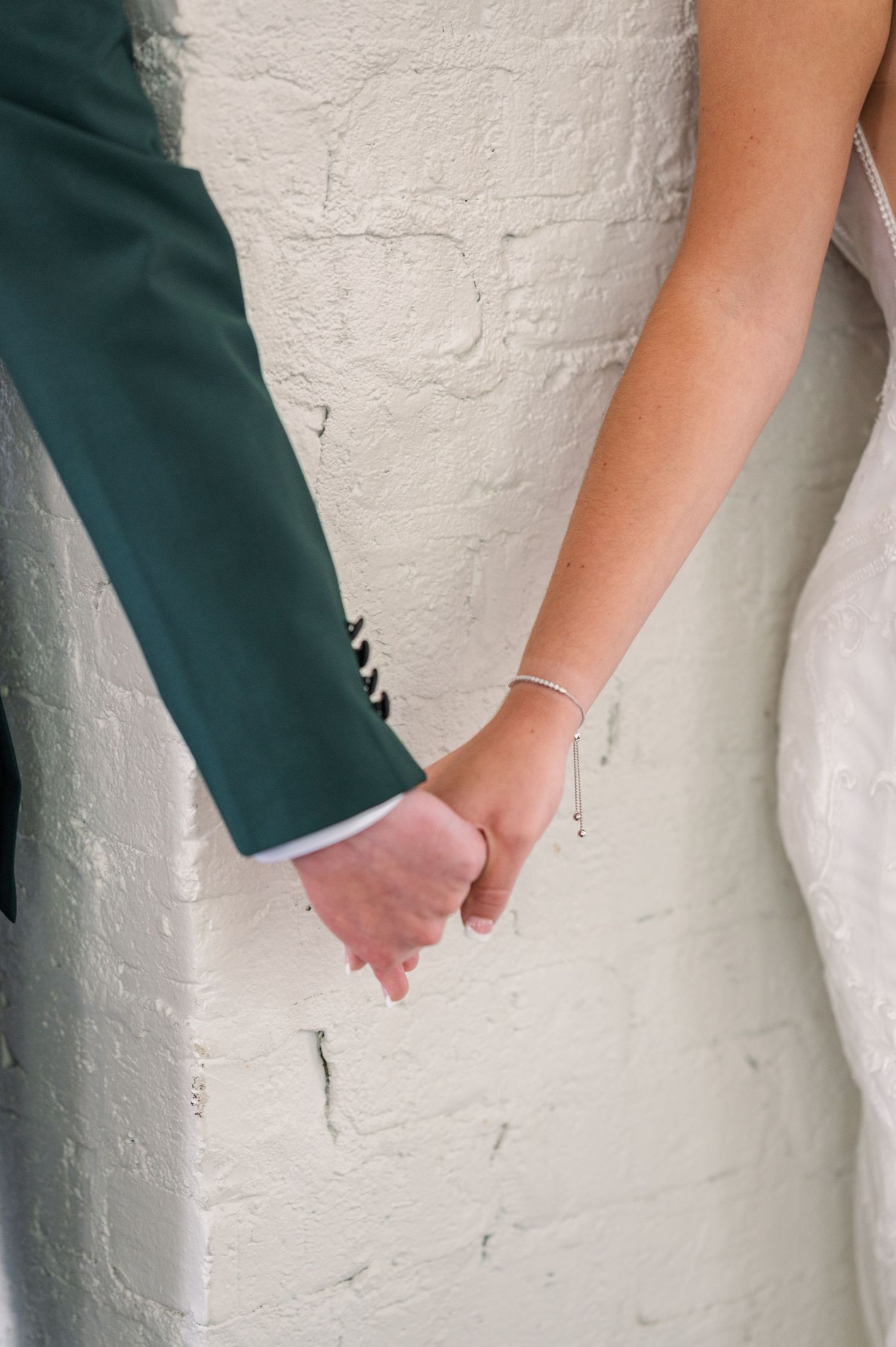 Close-up detail of hands holding during a wedding ceremony against a white textured wall.