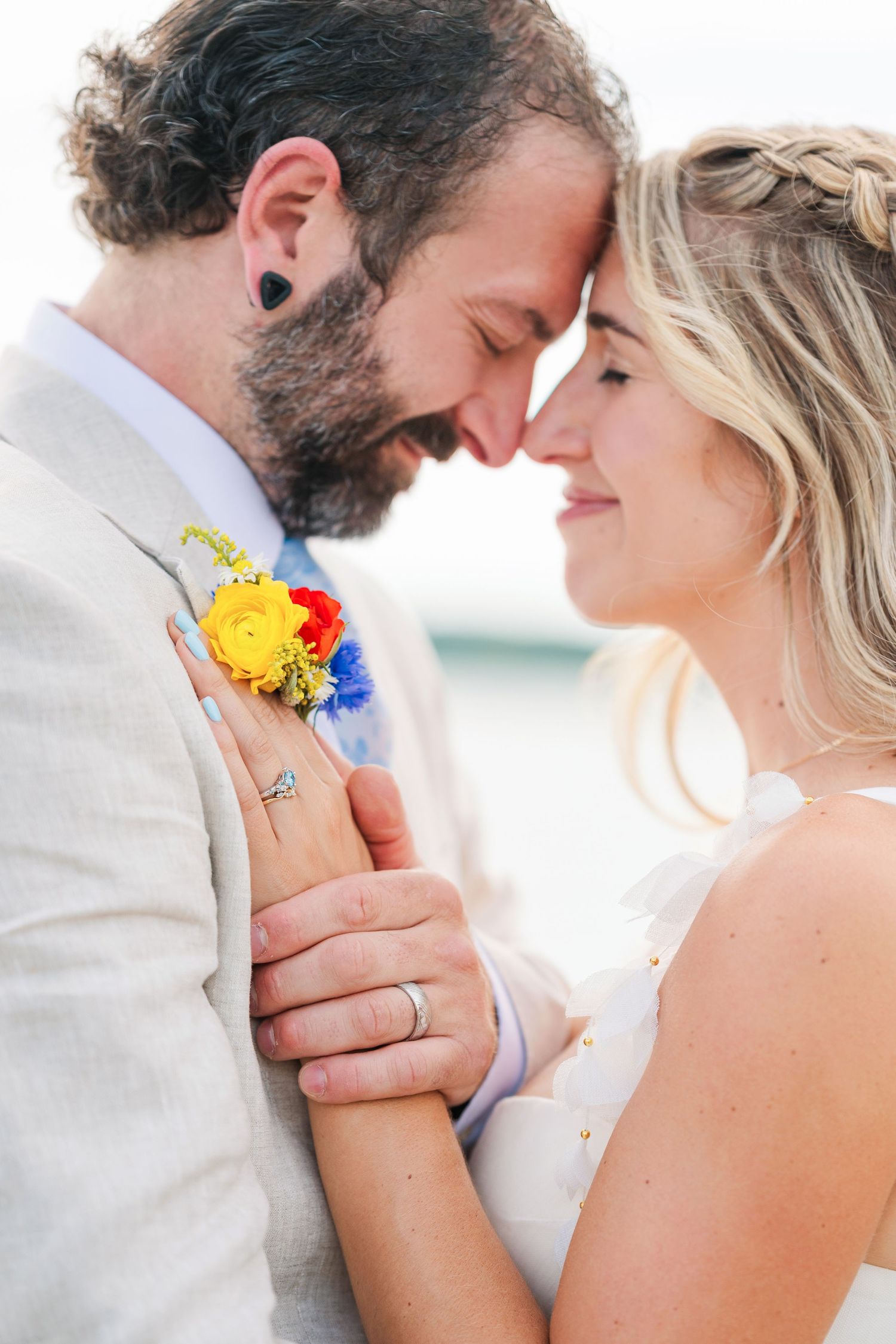 A bride and groom share an intimate forehead touch at their beach wedding, with colorful boutonniere and wedding rings visible.