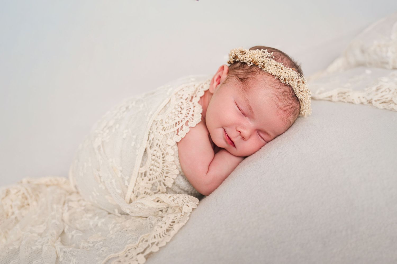 Smiling newborn baby wrapped in cream lace swaddle with floral headband, sleeping peacefully on soft white backdrop.