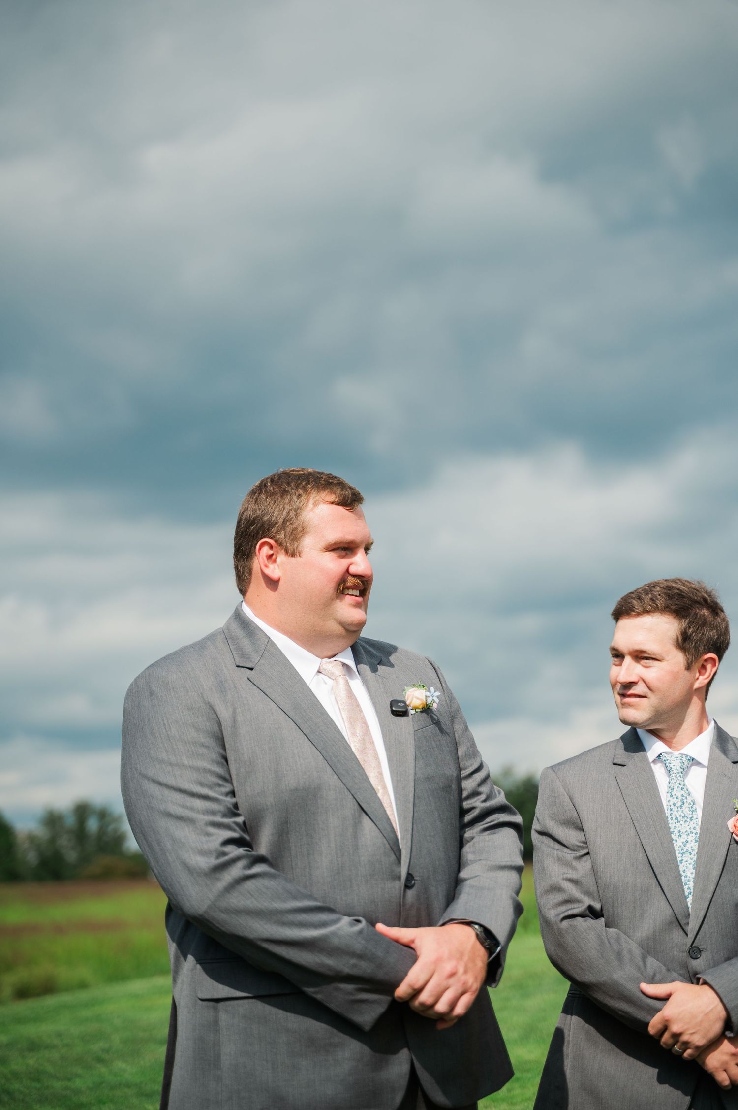 Two groomsmen in gray suits with boutonnieres smile outdoors at a wedding ceremony under a cloudy sky.