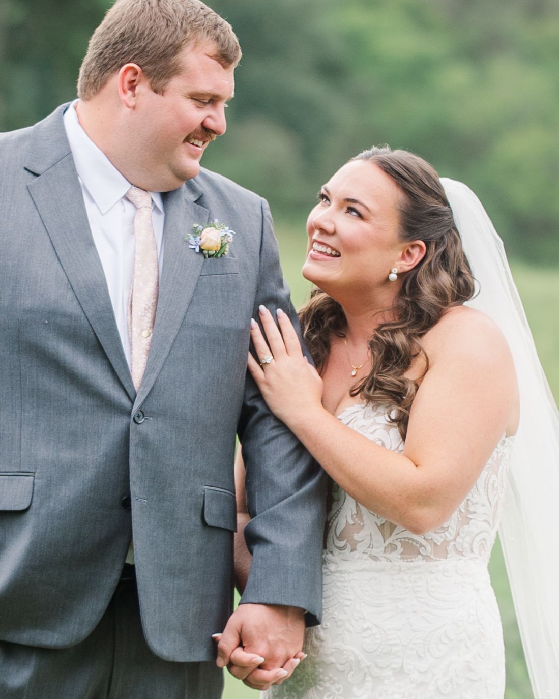 A smiling bride in a lace gown and veil holds hands with a groom in a gray suit with boutonniere outdoors.