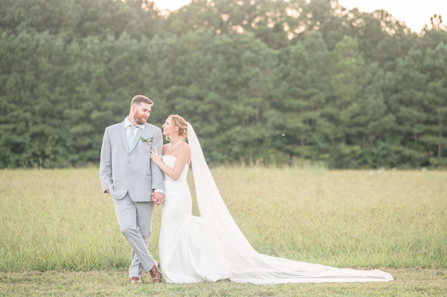 Elegant bride and groom pose in sunlit meadow with long flowing train.