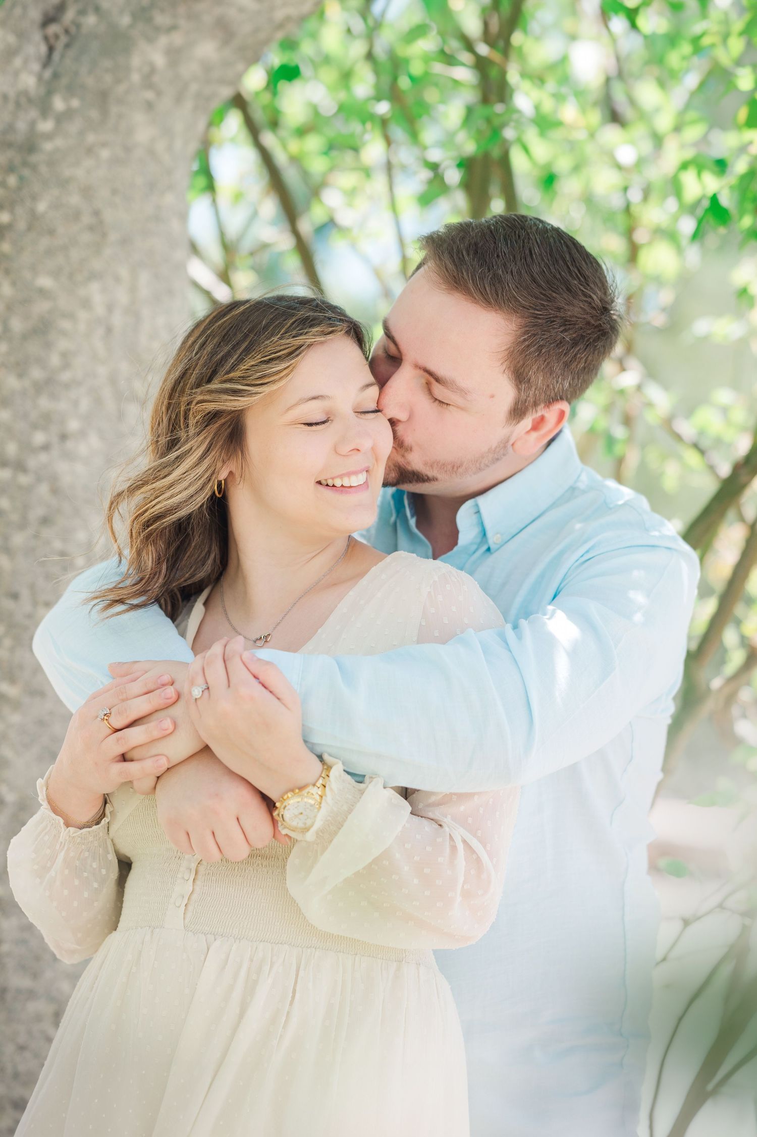 Intimate couple engagement session under green trees with soft natural lighting.