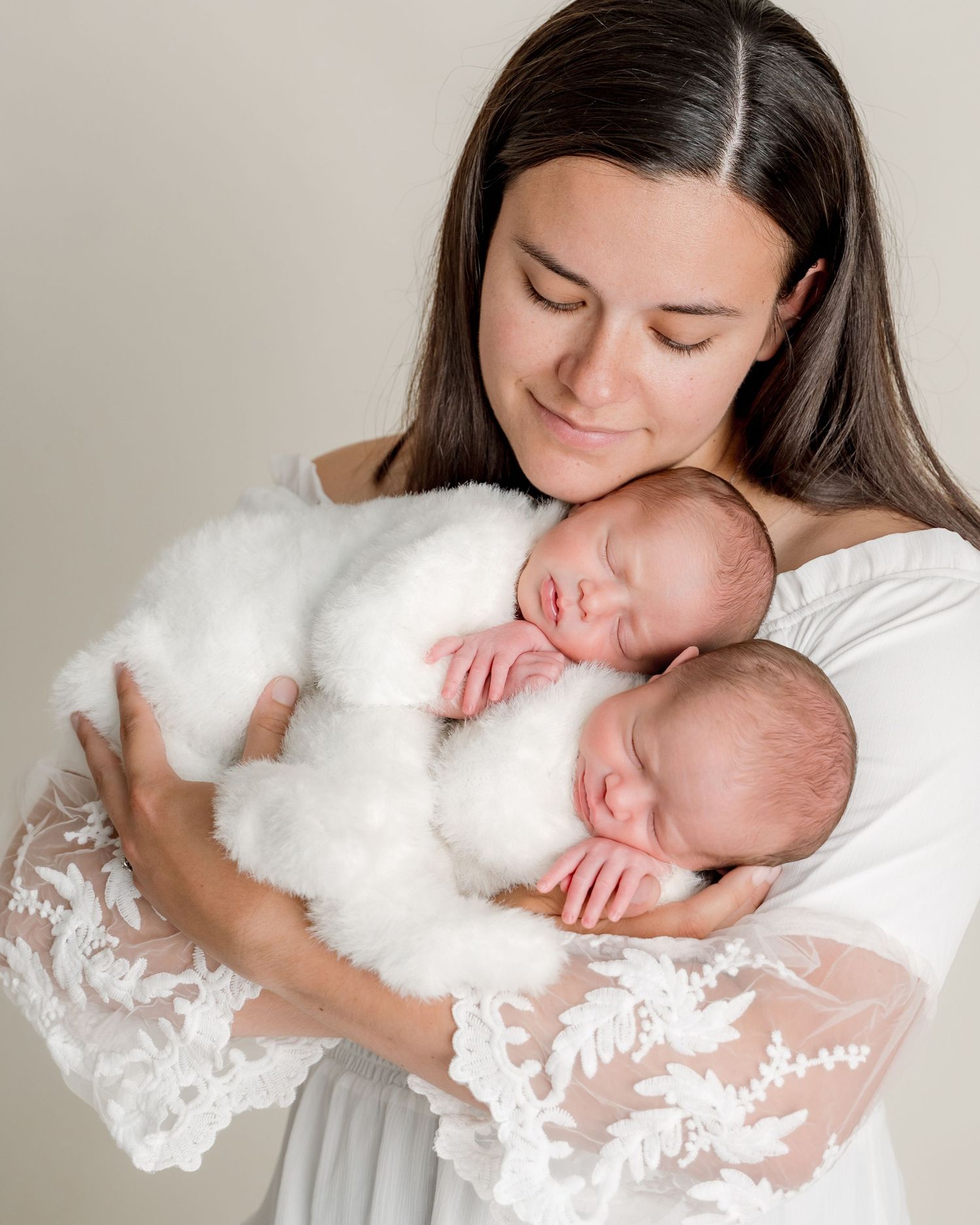 A smiling mother in a white lace gown tenderly holds her newborn twins in knit outfits.