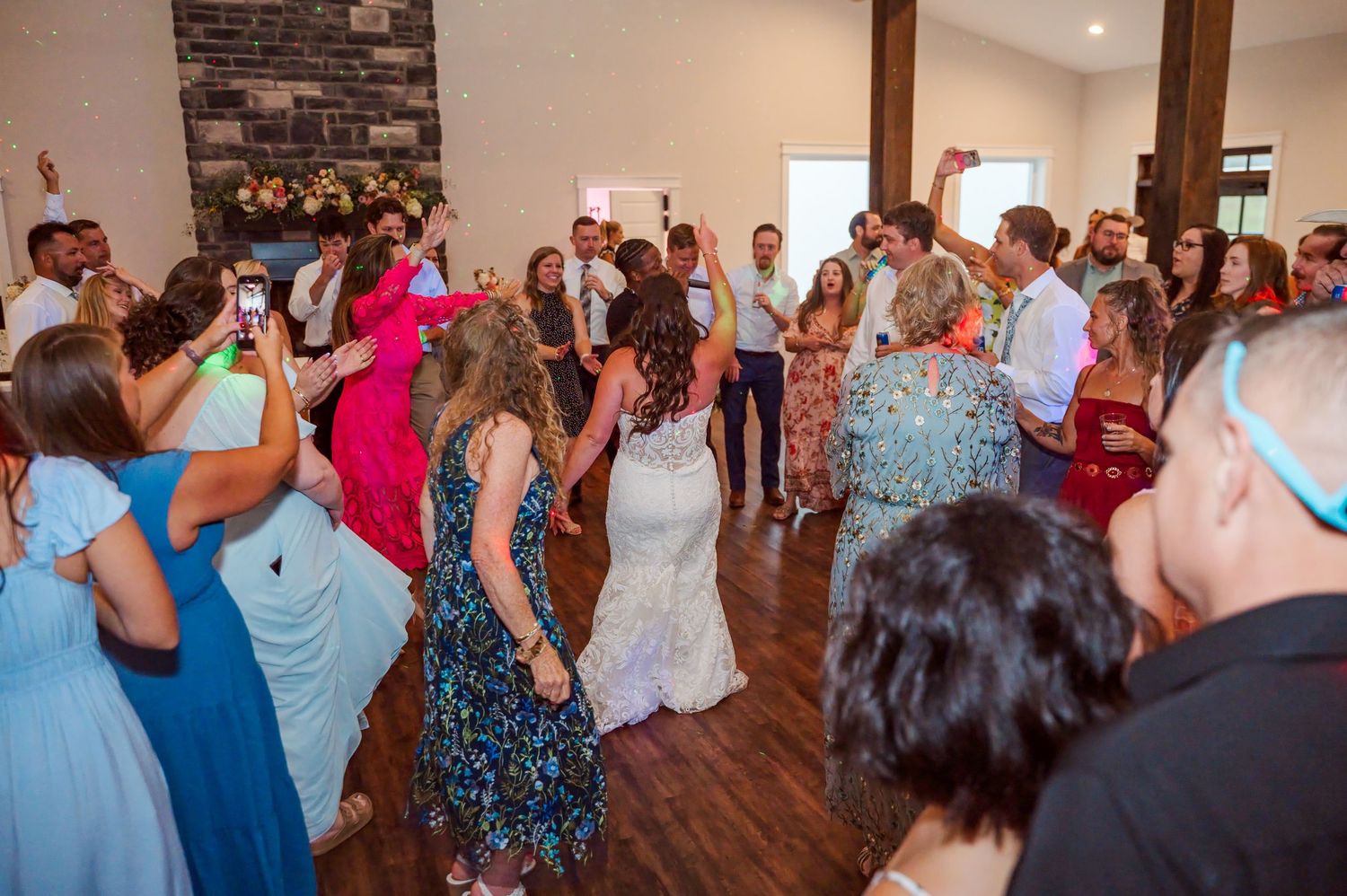 Wedding guests in colorful dresses dance together on a wood floor at a lively reception as a bride in white joins the celebration.