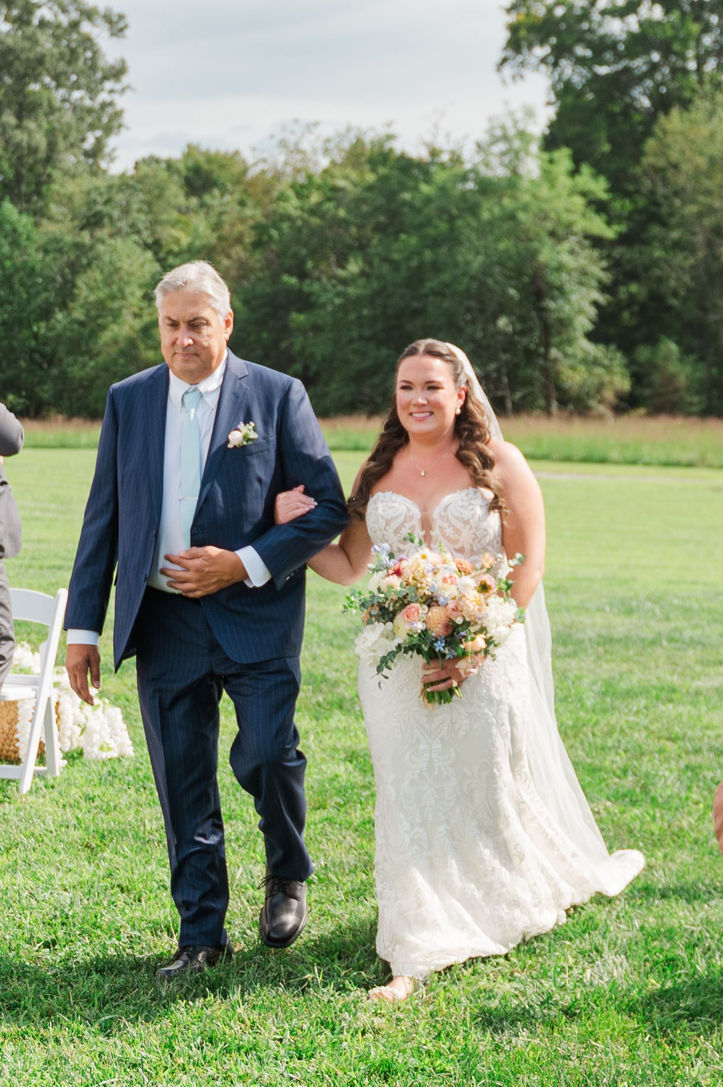 Bride in lace gown holding colorful bouquet walks down outdoor aisle with older man in navy suit.