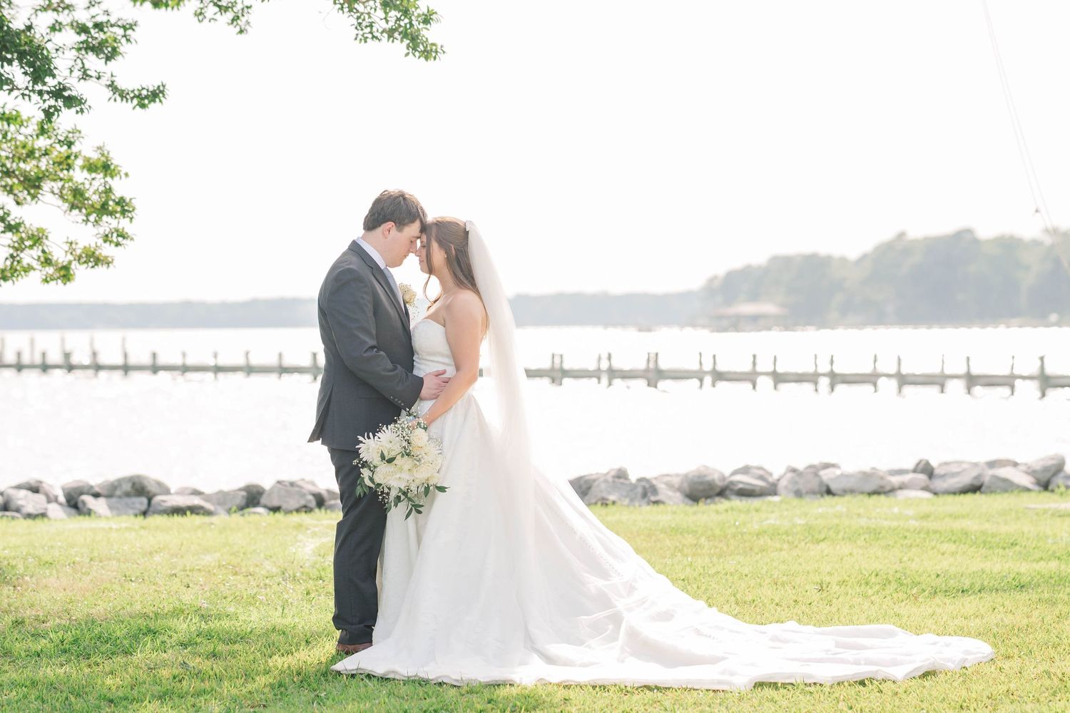 Newlyweds embrace on a grassy hill overlooking the water with a long white wedding dress trailing behind.