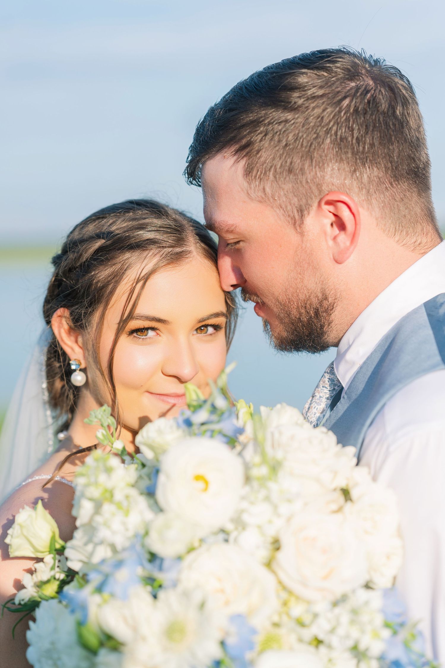 Bride and groom share a tender moment, bride holding white and blue bouquet, smiling softly outdoors on their wedding day.