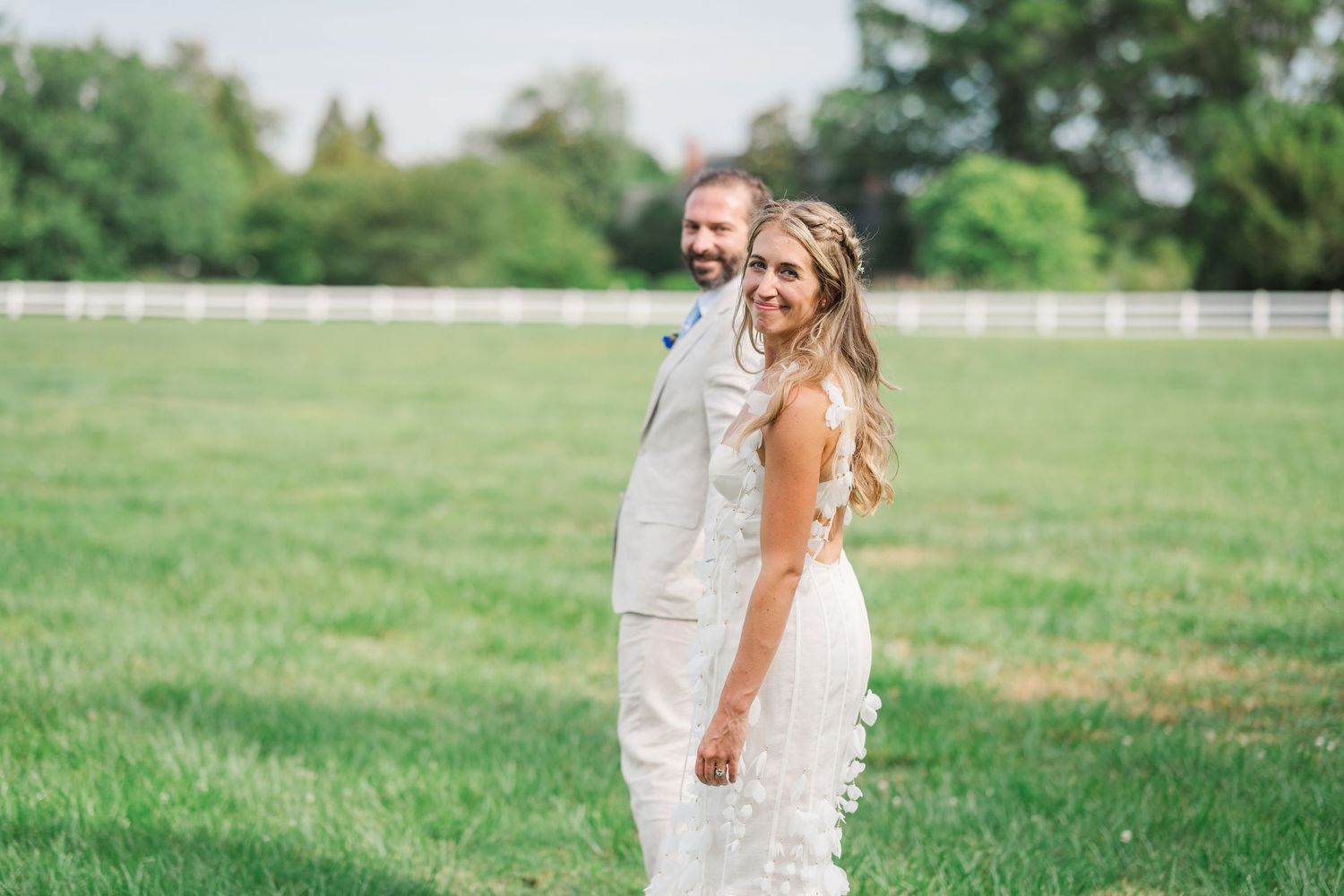 Bride and groom smiling outdoors on lush green lawn with white fence, bride in lace gown, groom in tan suit.
