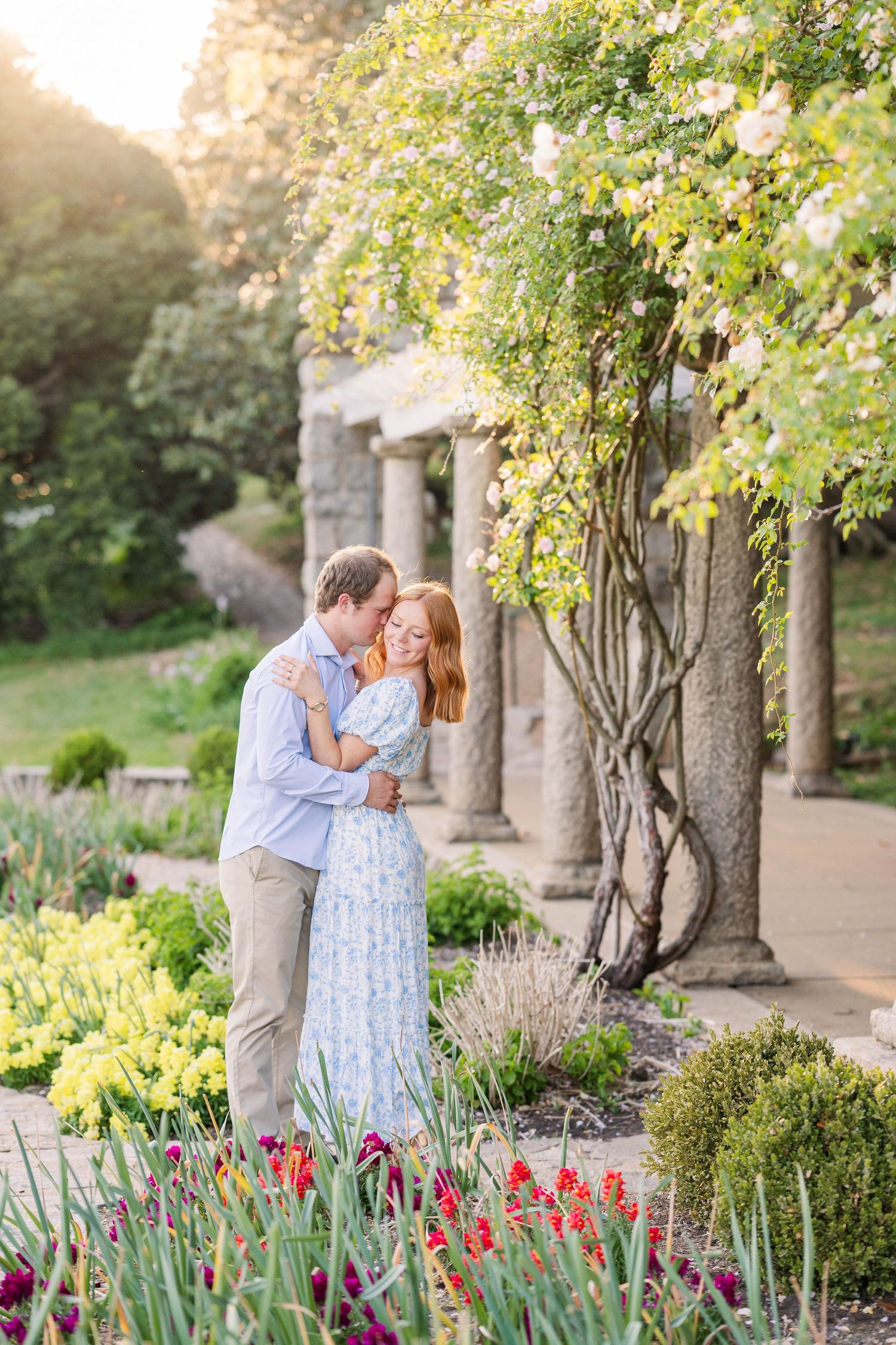 Couple embracing in a lush garden with colorful tulips, stone columns, and blooming trees in warm golden light.