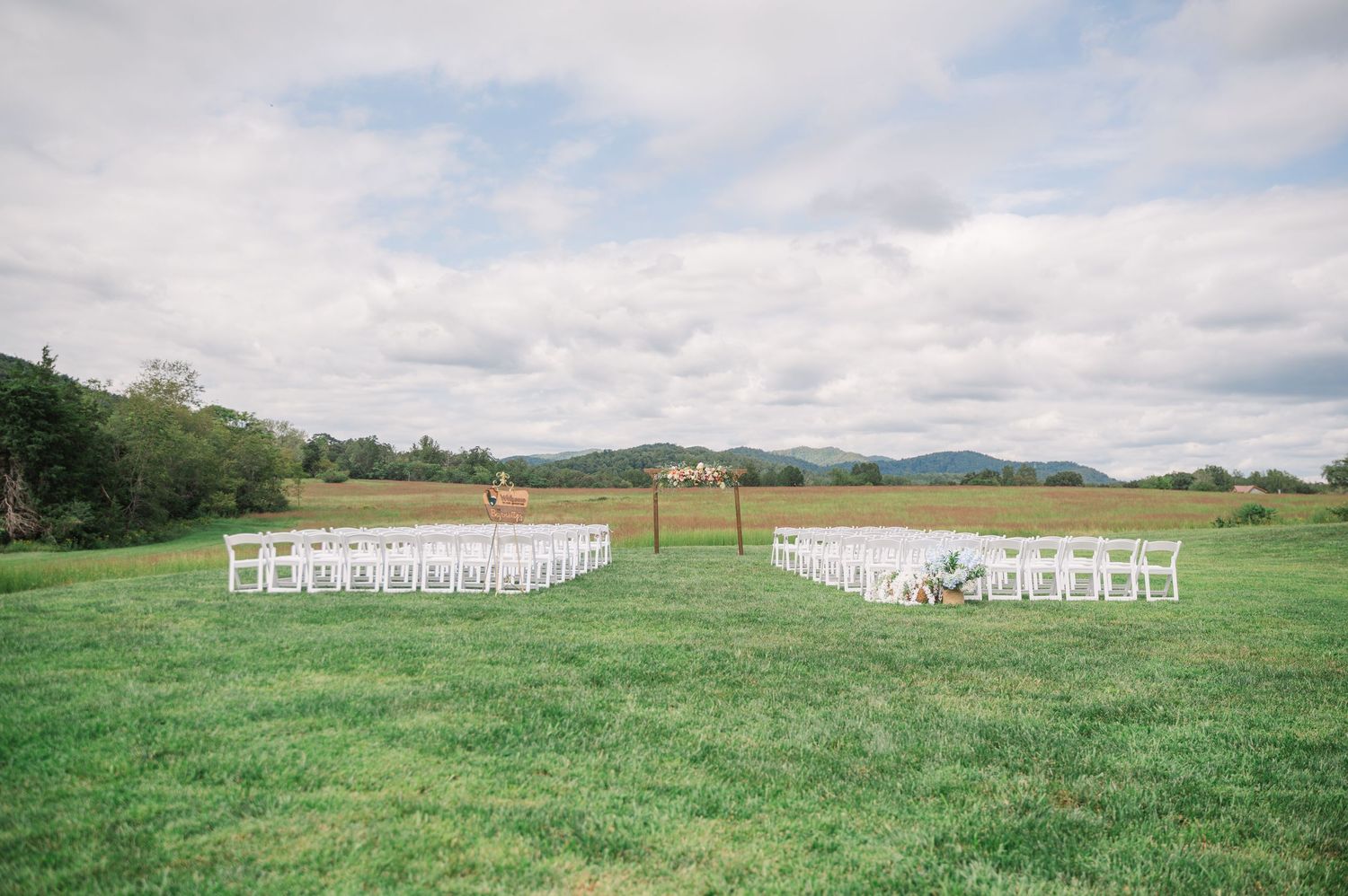 Outdoor wedding ceremony setup with white chairs on lush green grass, rolling hills and mountains in the background.