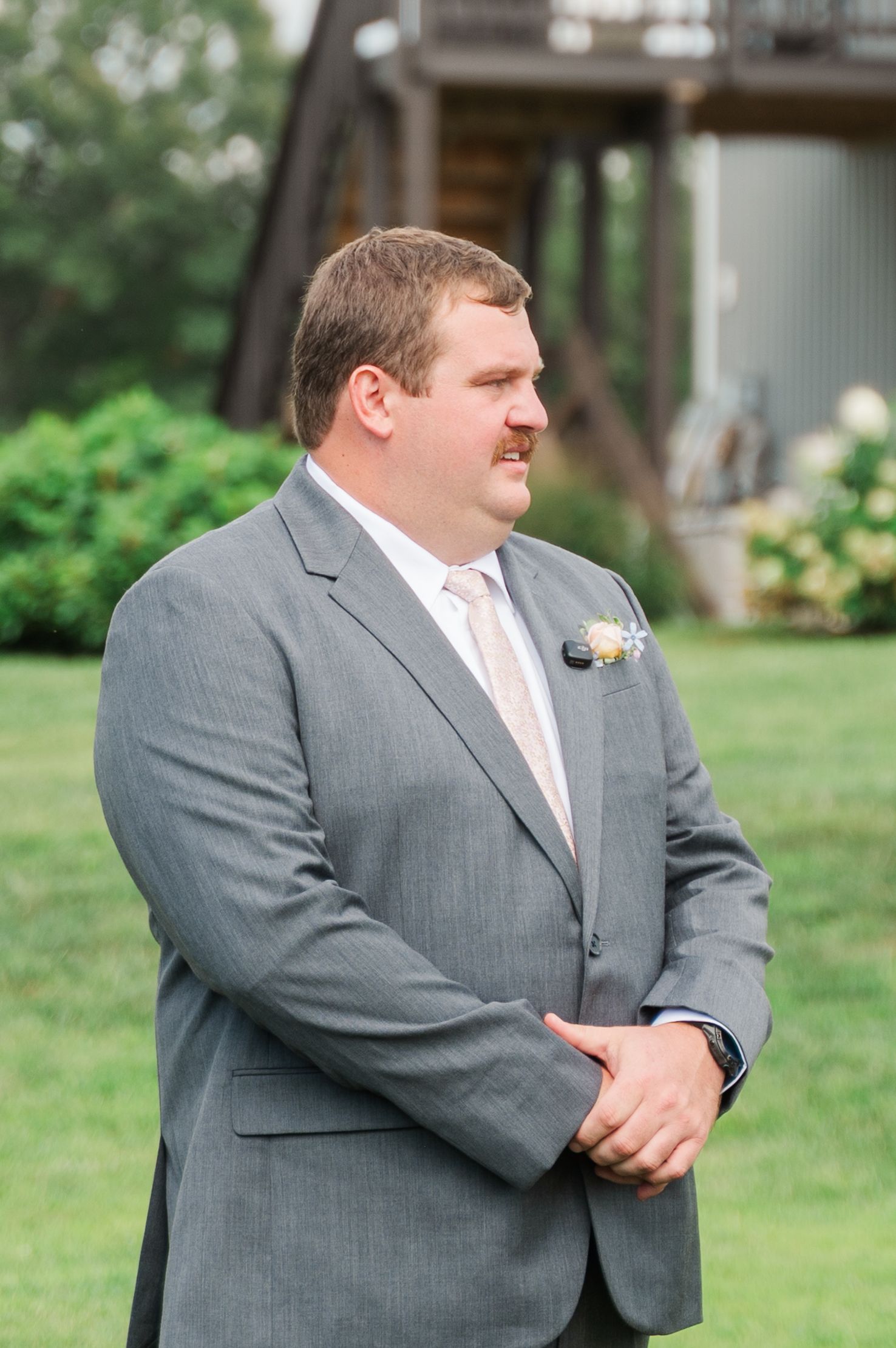Groom in gray suit with boutonniere smiling outdoors at wedding venue with green lawn and barn background.