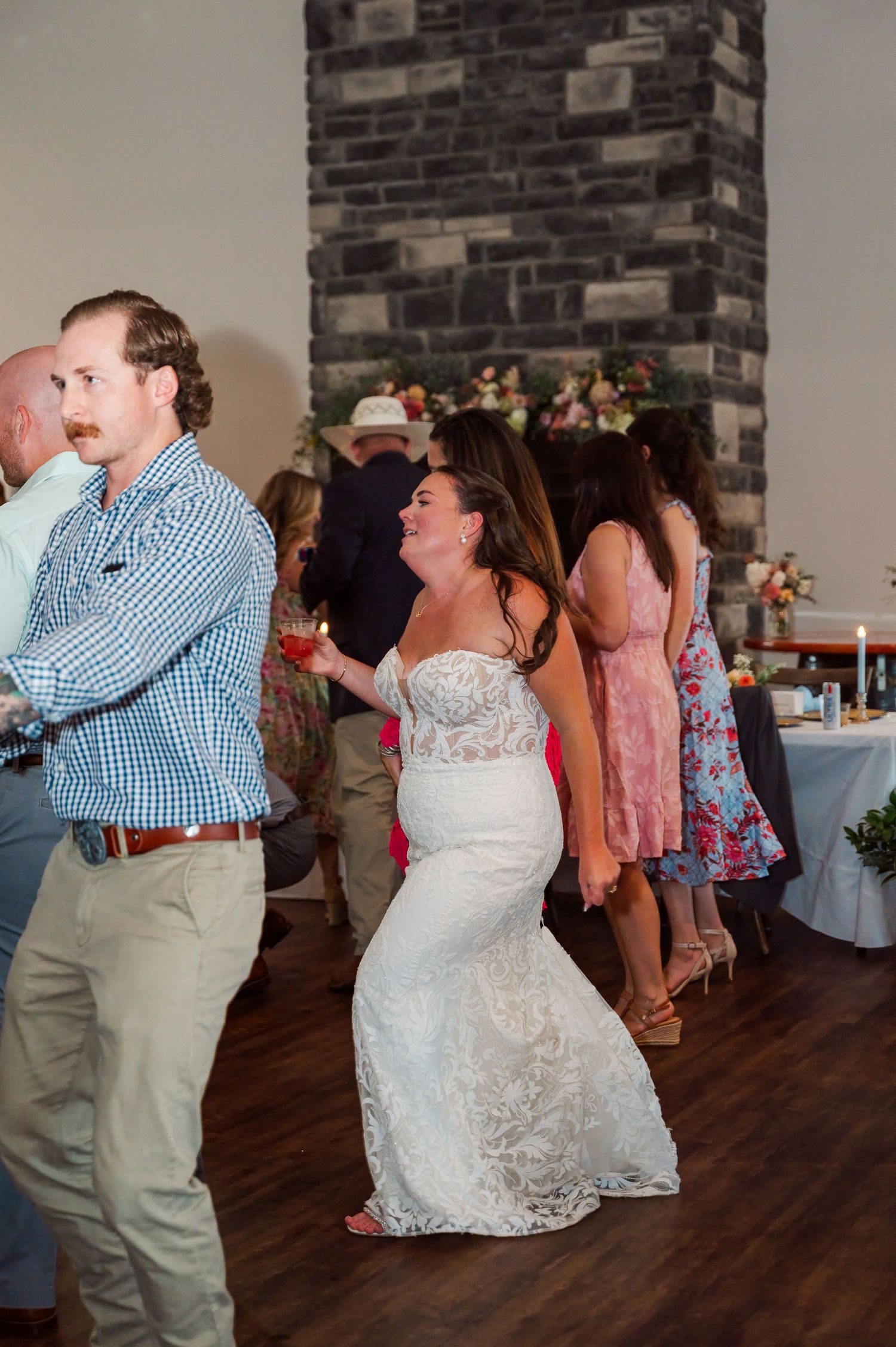 Bride in a lace wedding gown dances with guests on the wooden floor of a reception venue with a stone fireplace and floral decor.