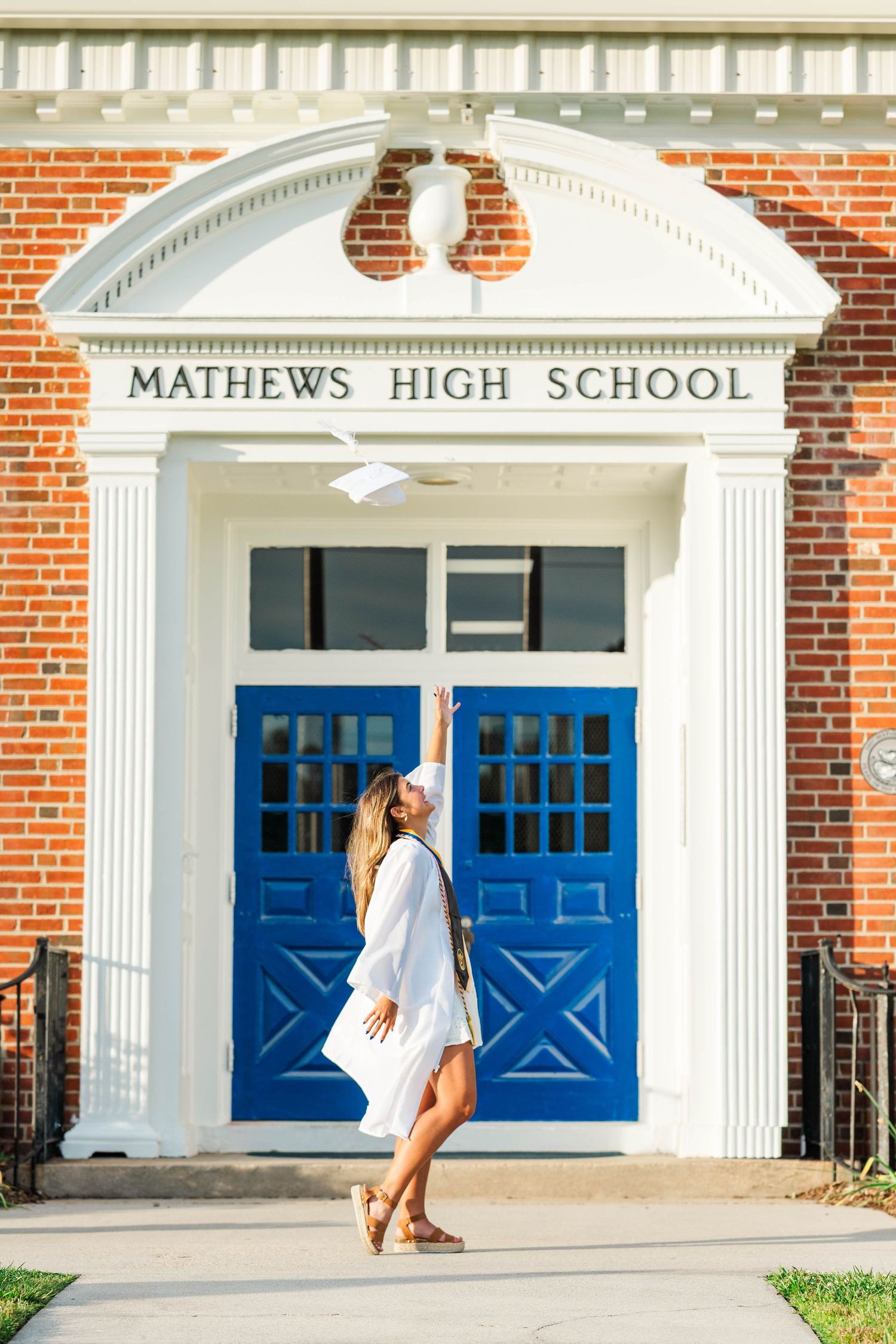 Graduate in white gown tosses cap in front of Mathews High School's iconic blue doors and brick facade.
