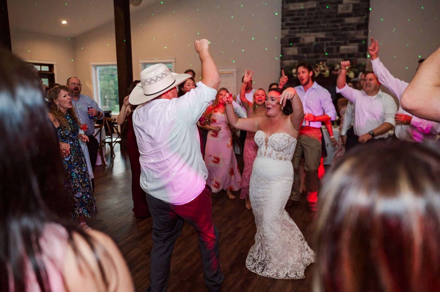 Bride in a white lace gown dances with a man in a cowboy hat as wedding guests cheer and clap around them on the reception dance floor.