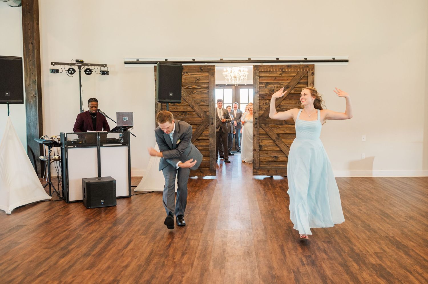 Wedding party members dance energetically on a hardwood floor inside a rustic barn reception venue with a DJ.