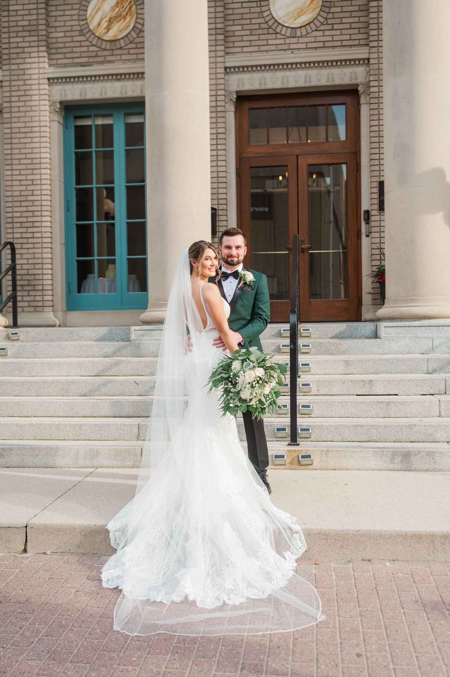 Elegant wedding couple poses on stone steps of historic post office building.
