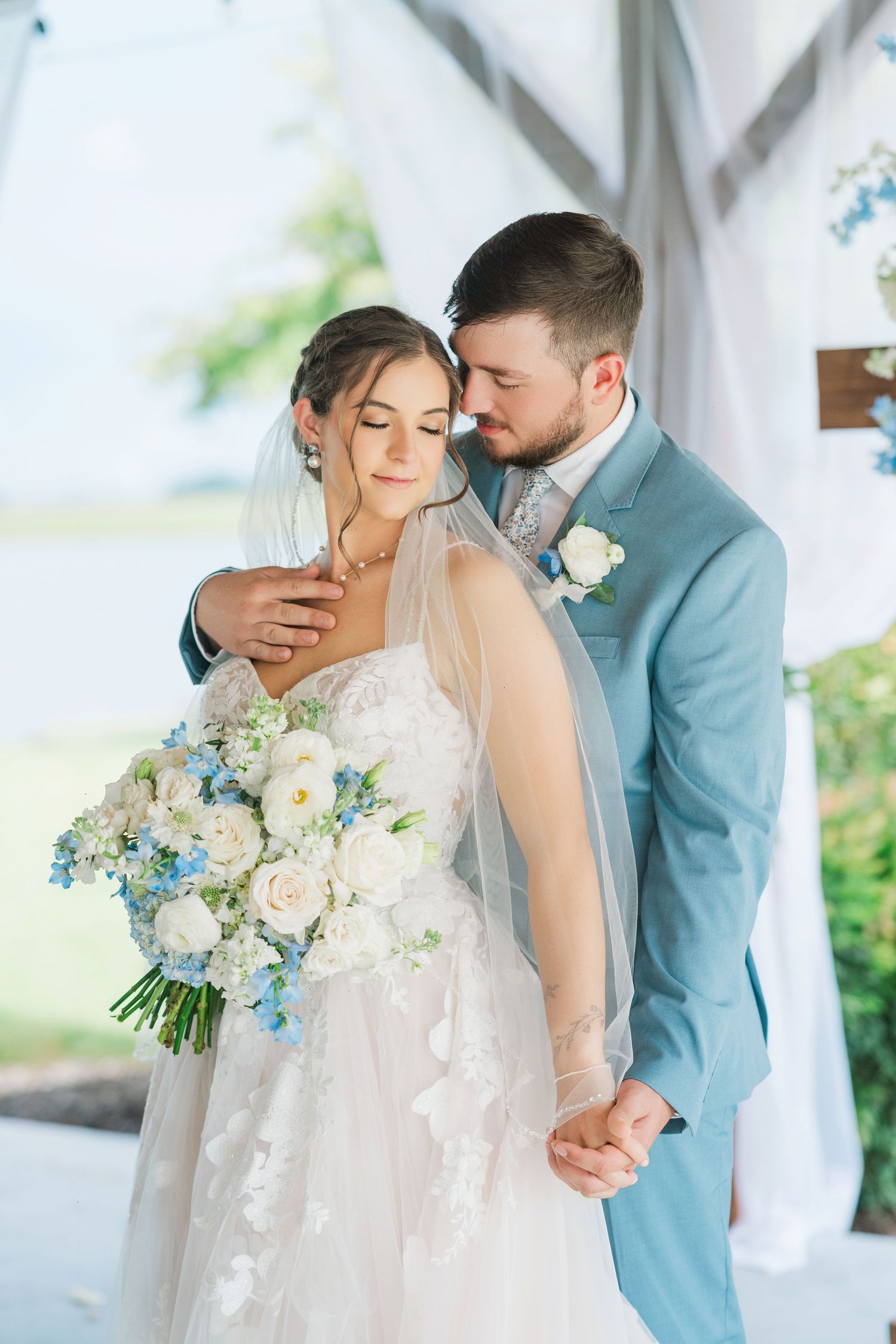 Bride and groom share a tender embrace, she holds white and blue bouquet, he wears light blue suit at outdoor wedding.
