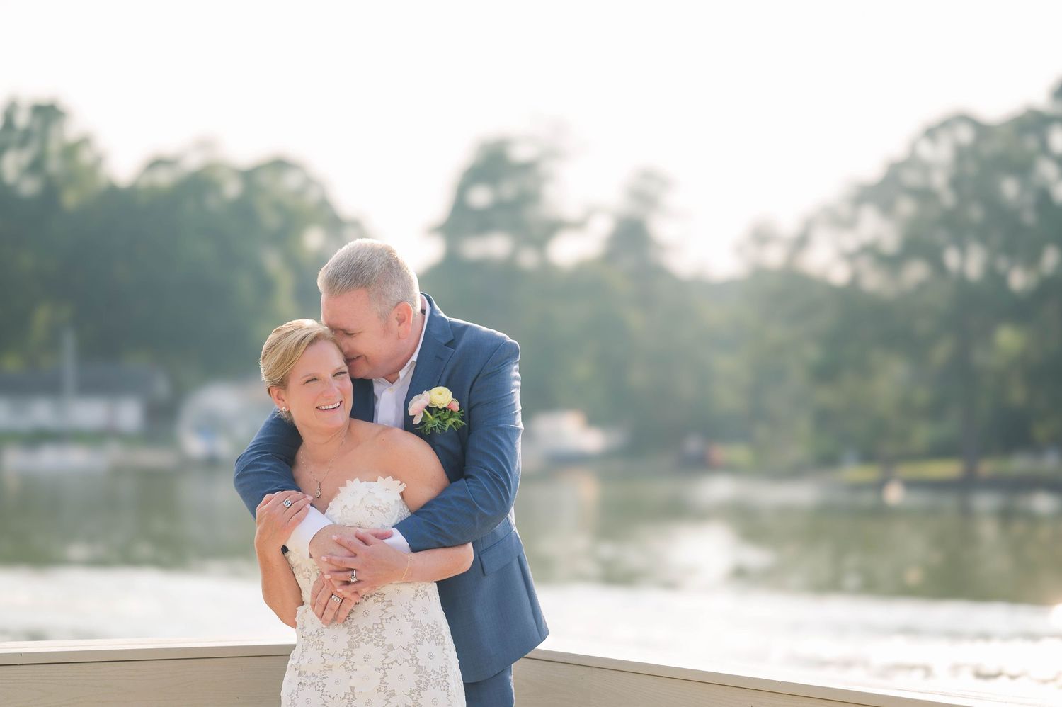Bride and groom embrace on a waterfront deck at golden hour, groom in blue suit kissing bride's cheek.