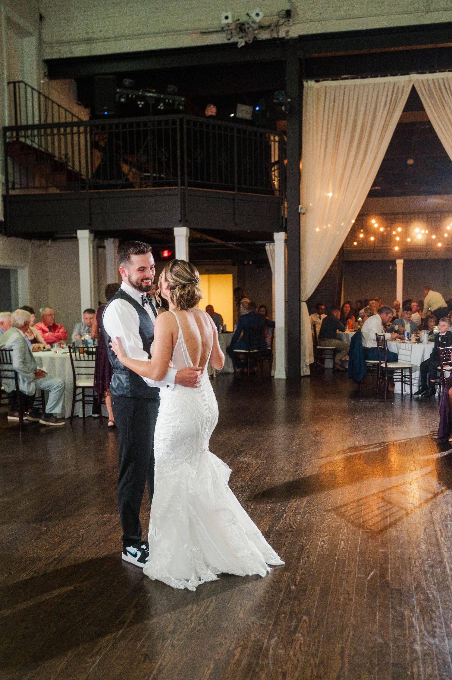 A couple dances on a dark wooden floor at their reception venue with string lights overhead.
