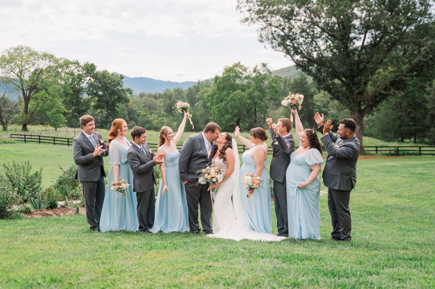 Wedding party celebrates outdoors on green lawn, bridesmaids in light blue dresses, groomsmen in gray suits, bride and groom kissing.