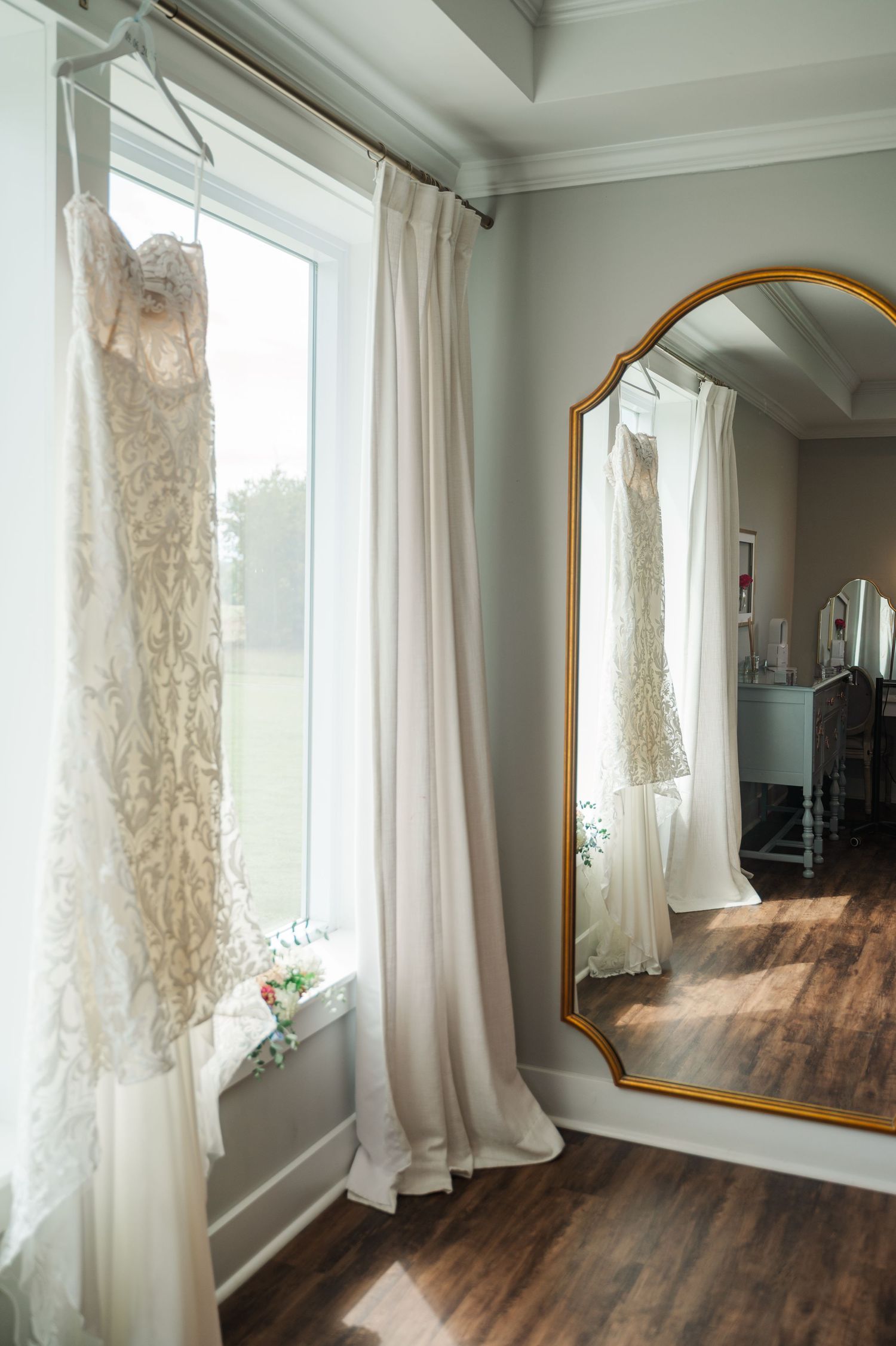 Elegant lace wedding dress hanging by a bright window reflected in a gold-framed floor mirror.