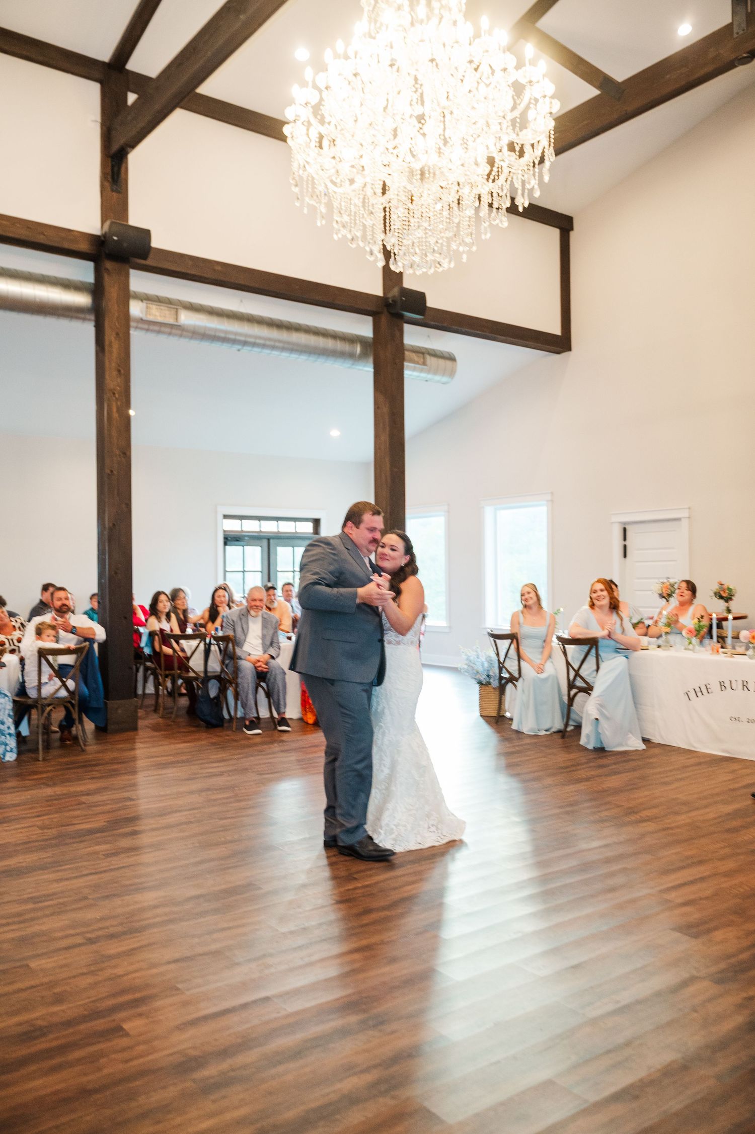 Bride and groom share a first dance on a wooden floor beneath a crystal chandelier as wedding guests watch from tables.