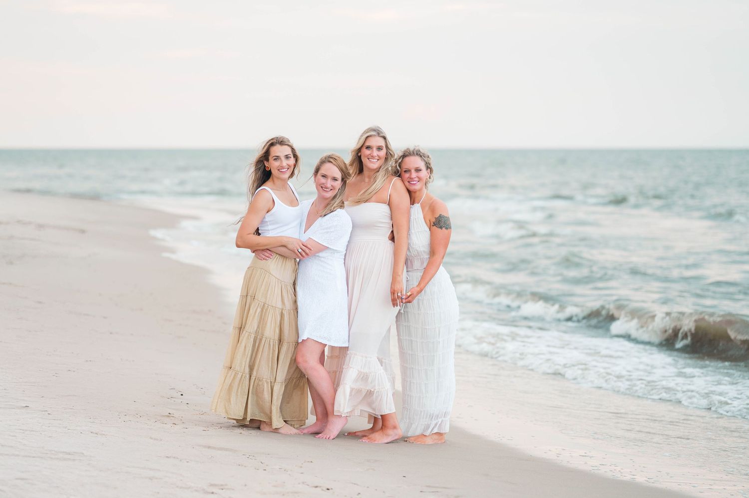 A group of friends in light-colored dresses pose together on a serene beach at sunset with ocean waves in the background.