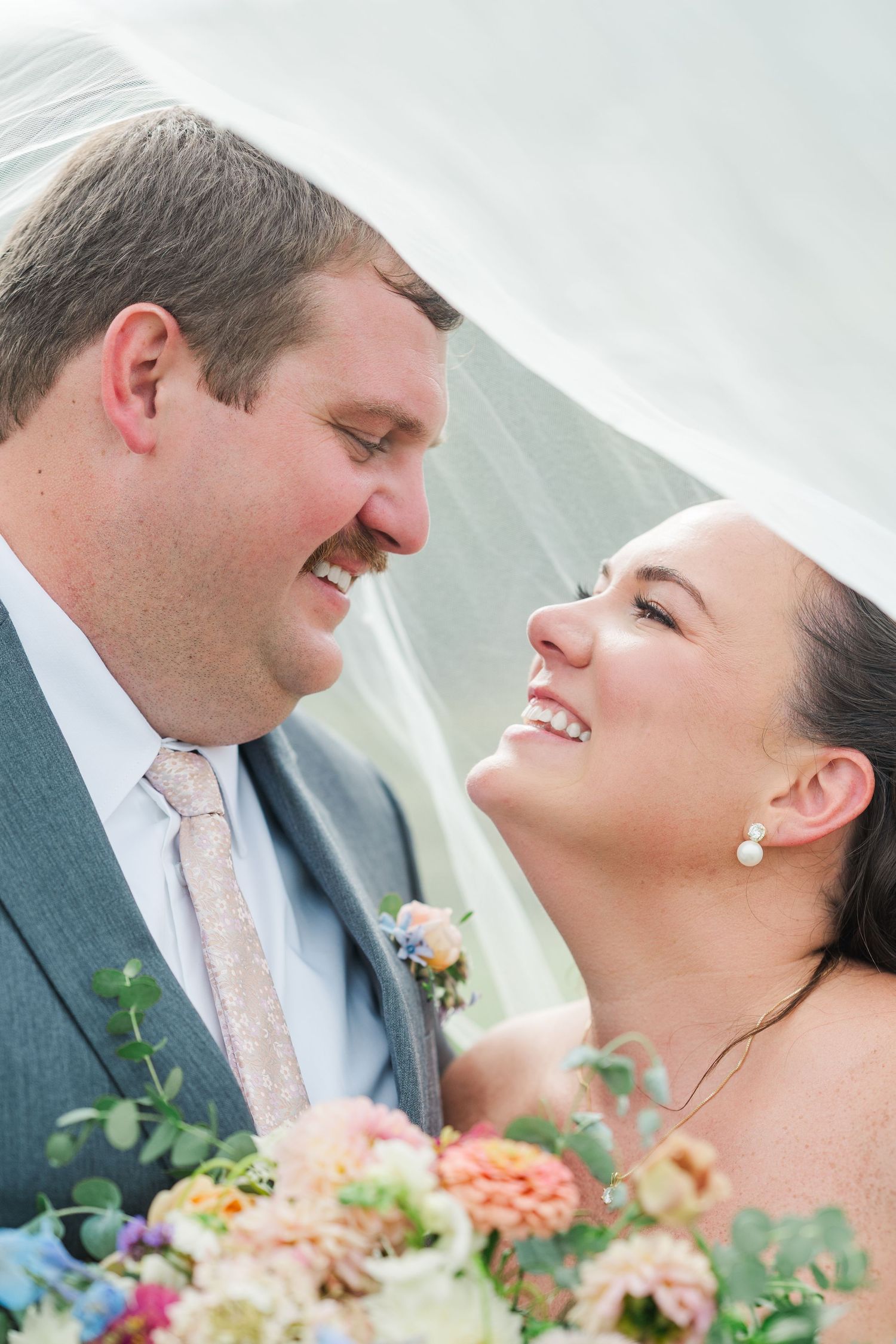 Bride and groom smiling at each other under a wedding veil with a colorful bridal bouquet.