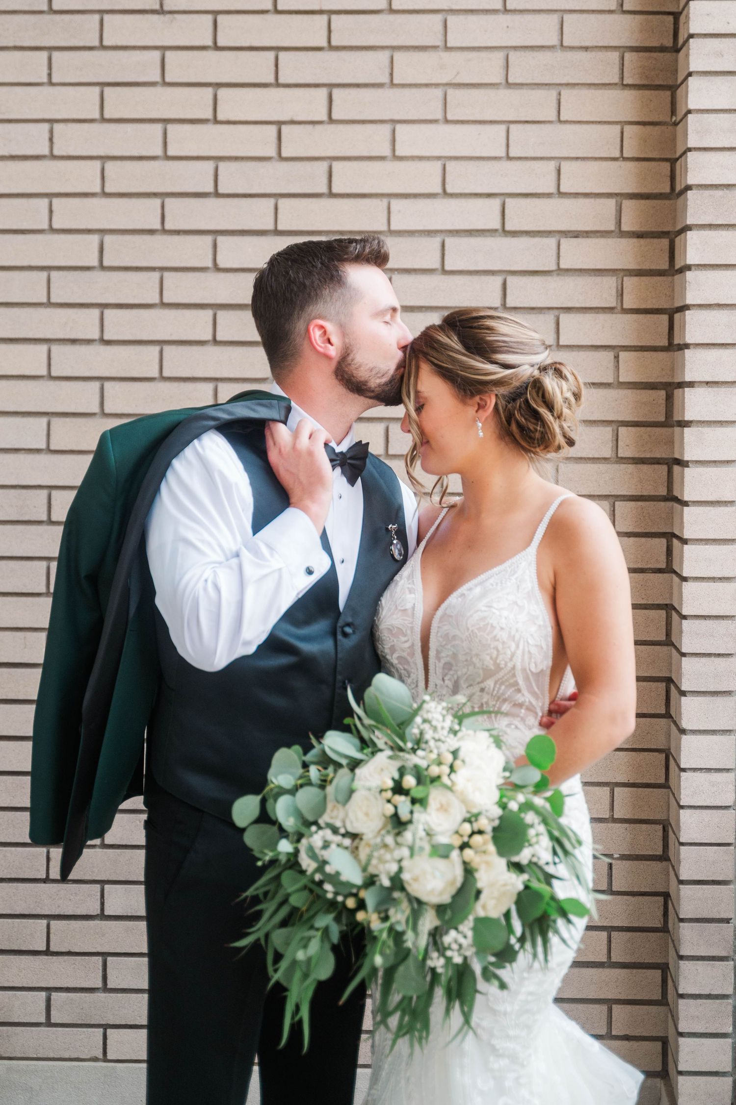A romantic wedding moment captured against a light brick wall with elegant greenery bouquet.