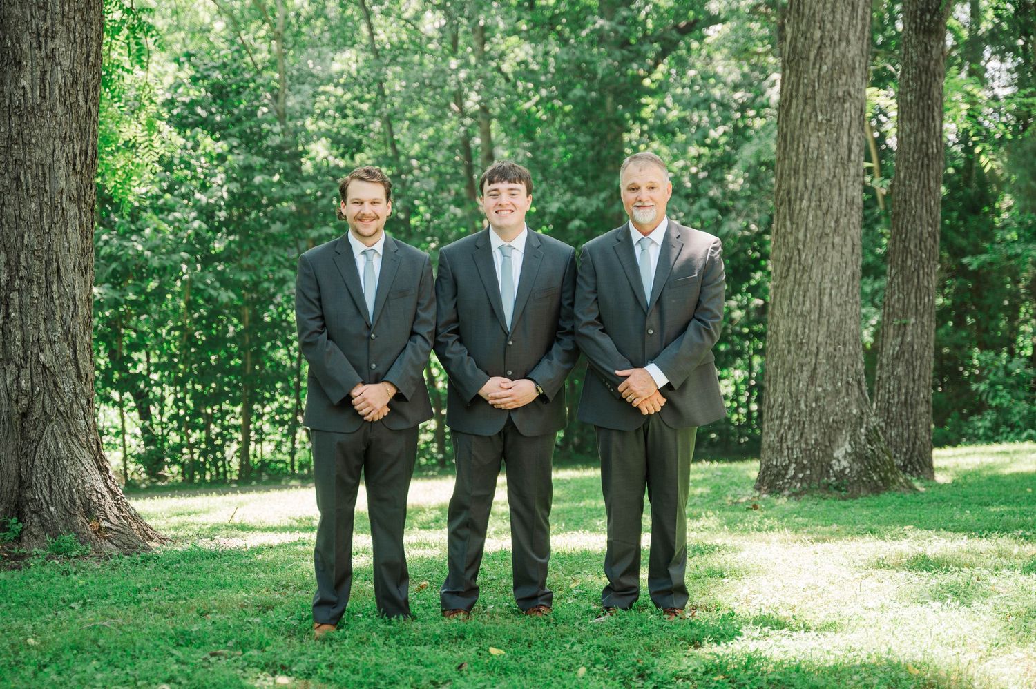 Small group in formal attire standing together between trees outdoors.