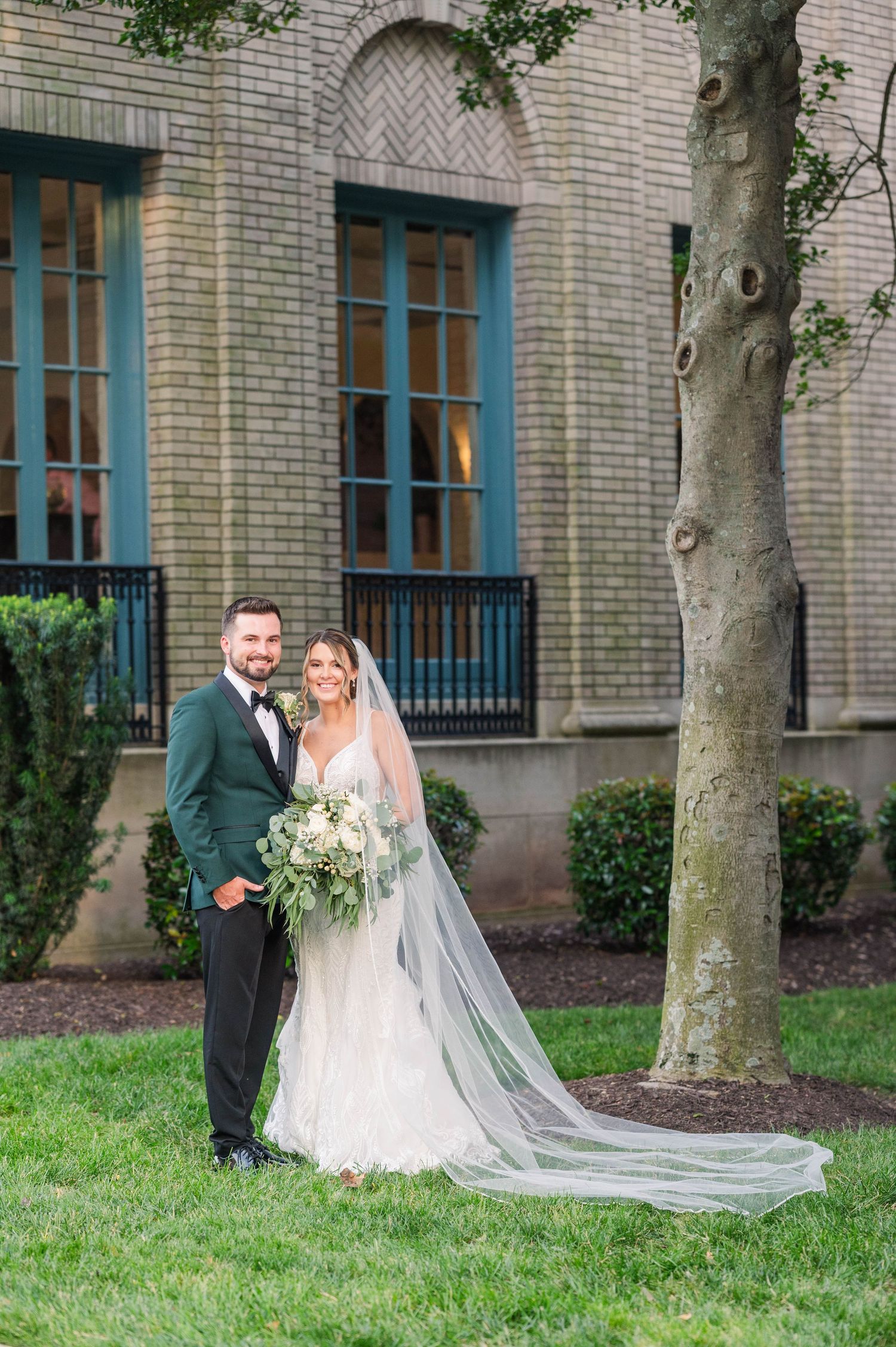 A couple poses on a lawn with trees and a long flowing veil near a brick building.