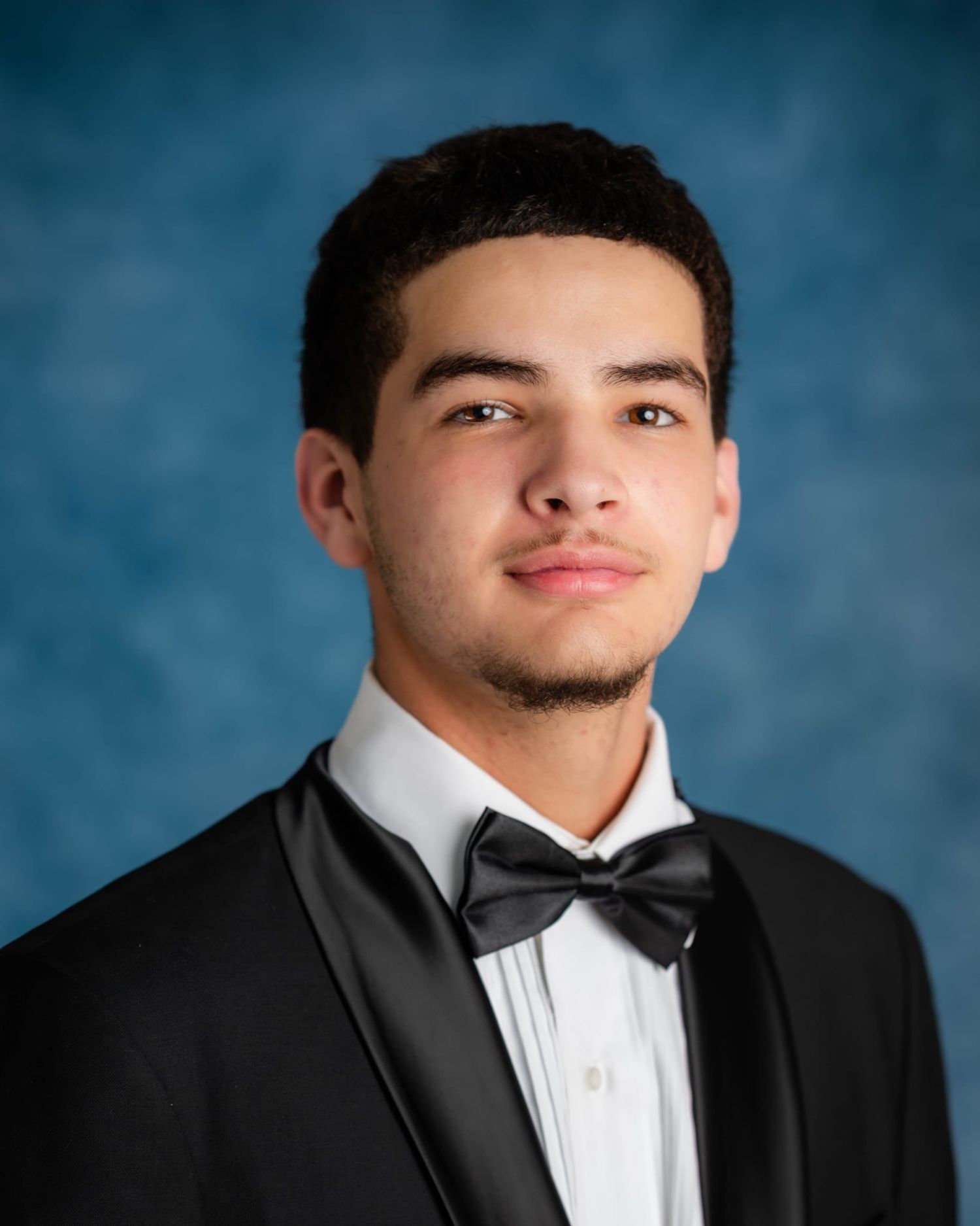 Young man in black tuxedo with bow tie poses for formal portrait against blue background.