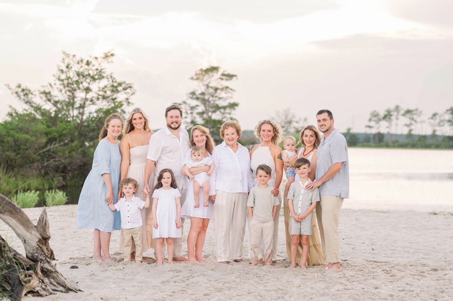 A group in coordinated light blue and white outfits poses together on a beach at sunset with water and trees in background.