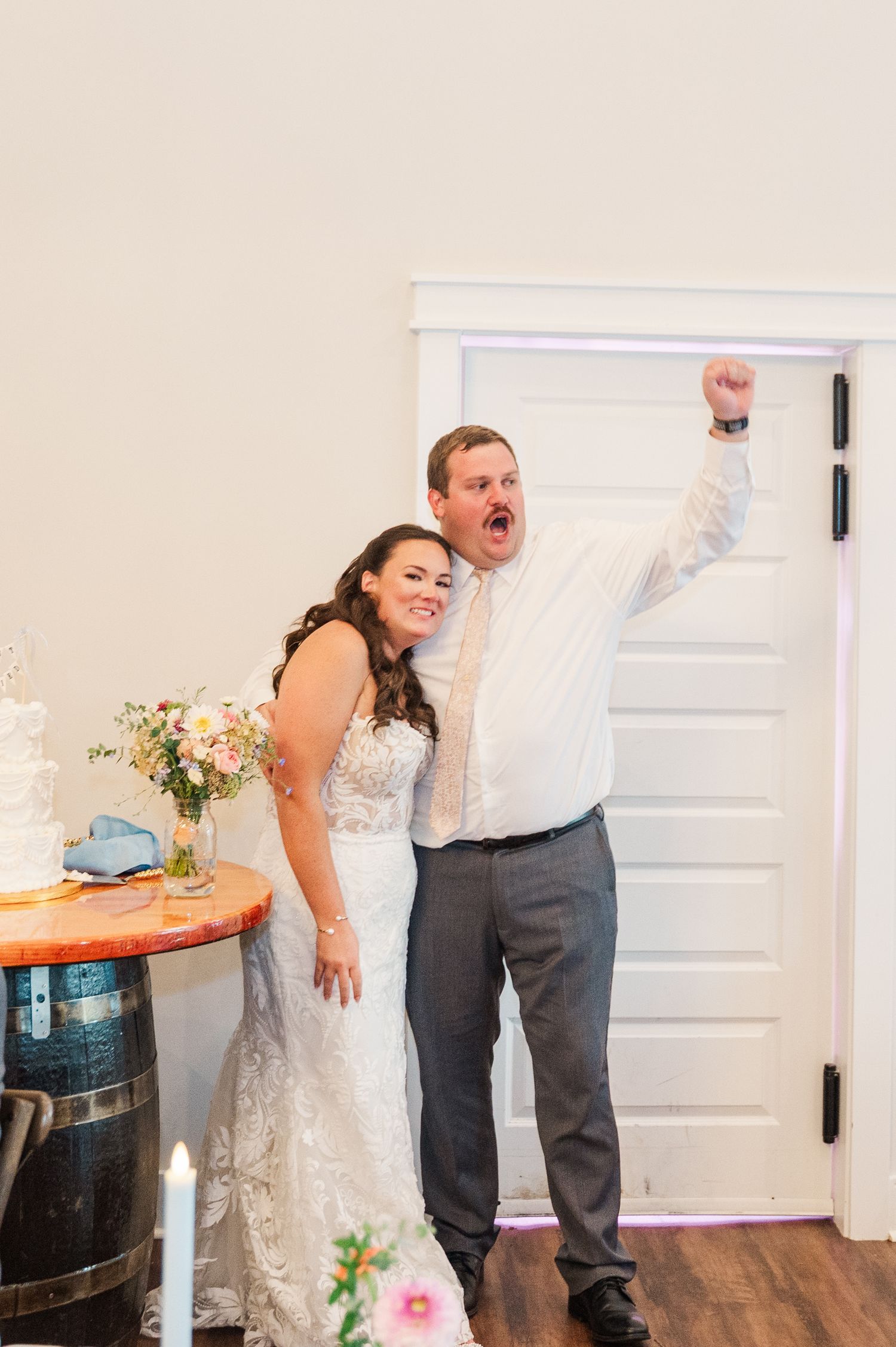 Bride and groom celebrating joyfully near wedding cake, groom raising fist in excitement at indoor reception venue.