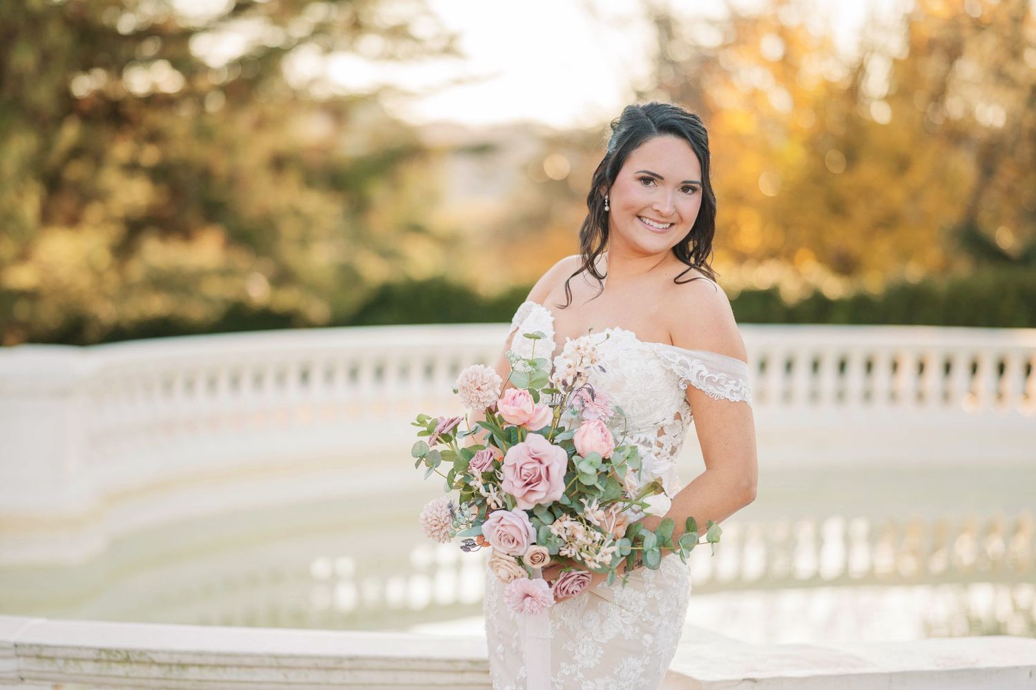 Bride in off-shoulder lace gown holds pink floral bouquet outdoors at golden hour near elegant stone fountain.