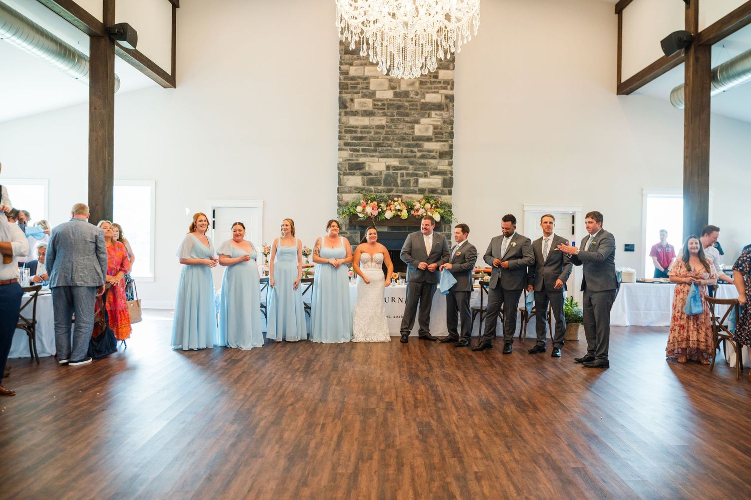 Wedding ceremony in elegant venue with chandelier, stone fireplace, bridesmaids in blue dresses, and guests gathered on hardwood floors.