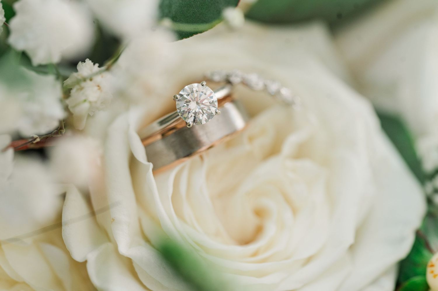 wedding bands and engagement ring on white rose bouquet.