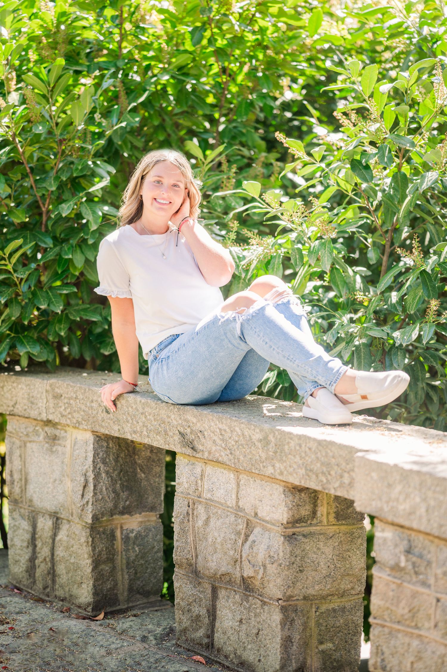 Casual portrait of person sitting on stone wall with green foliage background.