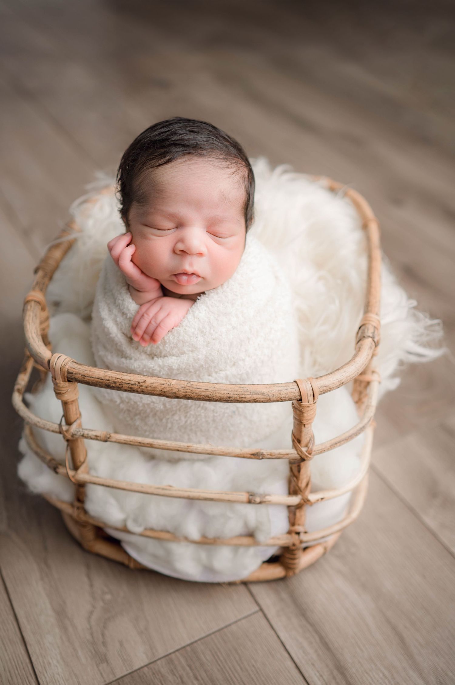 Sleeping newborn wrapped in soft white blanket nestled in rustic woven basket on wooden floor.