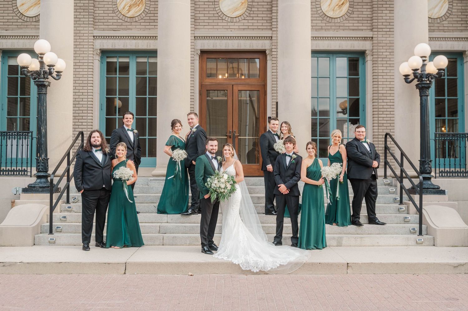 Wedding party gathered outside grand brick building with columns and ornate architecture.