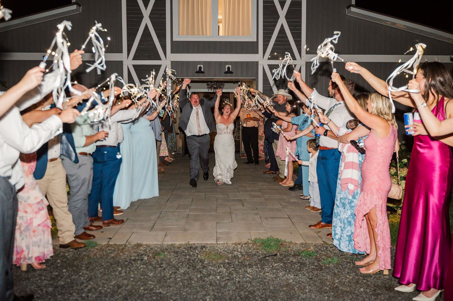 Wedding guests form a sparkler send-off tunnel as the bride and groom walk hand in hand outside a rustic barn venue.