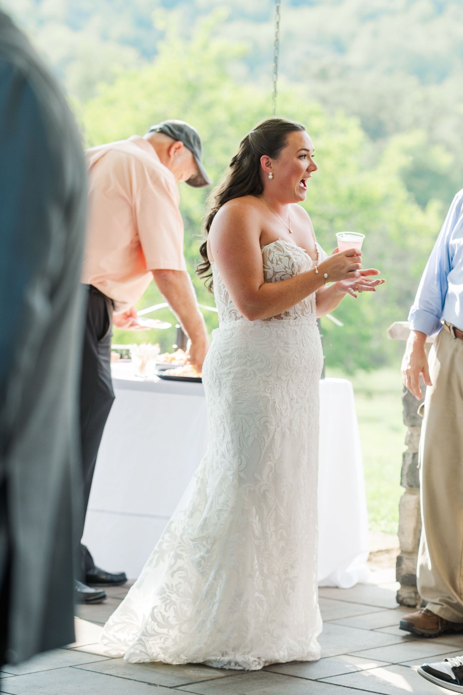 Bride in lace wedding gown laughing joyfully at outdoor reception with scenic green hills in background.